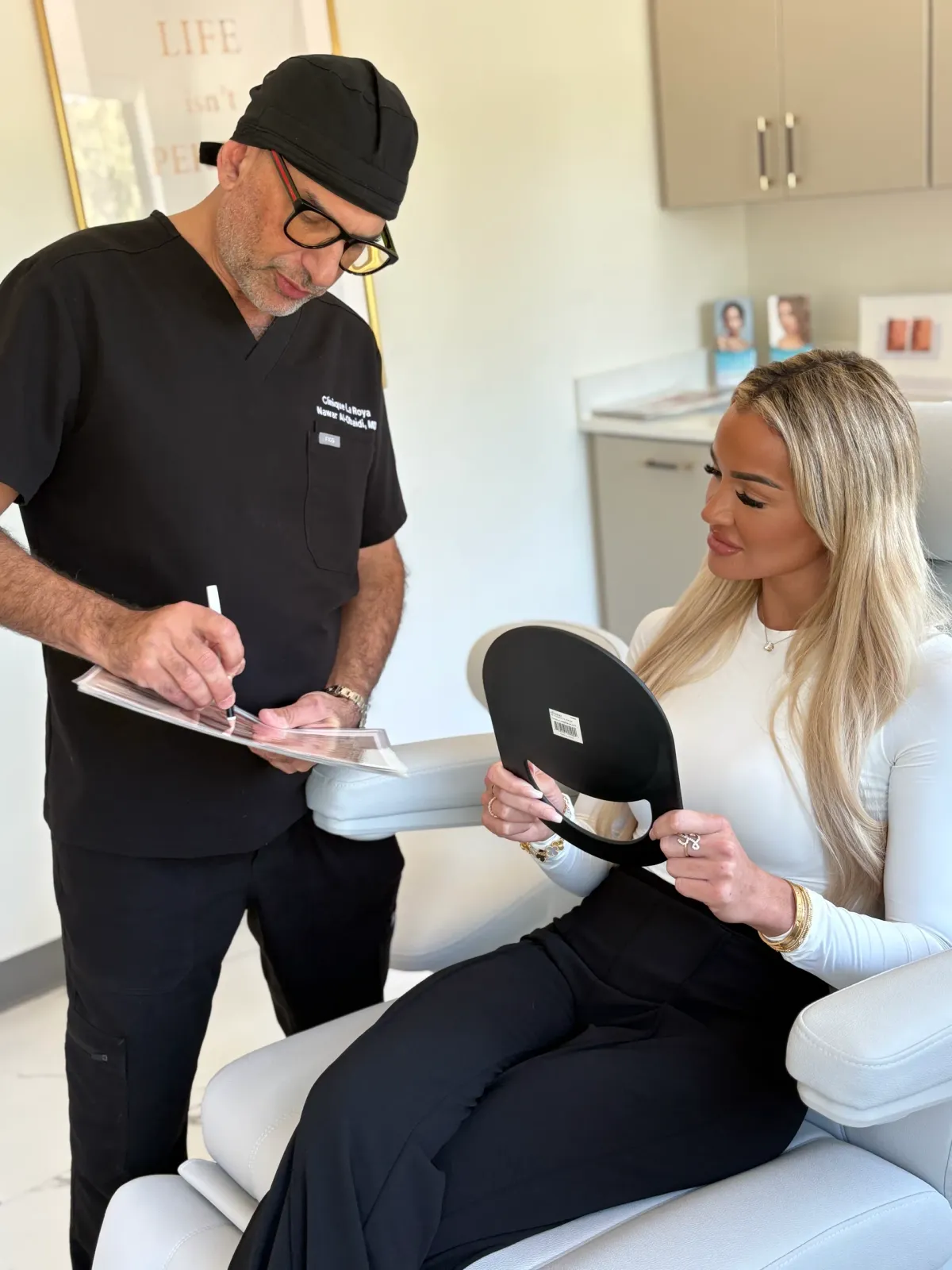 Dr. Nawar Al-Obaidi noting a filler plan while a patient studies her face in a hand mirror